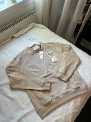 Beige sweatshirt från Ami Paris - Säljer en beige sweatshirt från Ami Paris med deras ikoniska hjärtlogga broderad på bröstet. Tröjan har rund hals, ribbade muddar och är gjord i mjukt bomullsmaterial. Perfekt för en chill och stilren look.