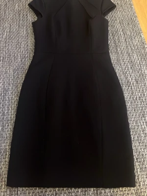 Svart kortärmad klänning med krage - Stilren svart klänning med korta ärmar och elegant krage. Klänningen har figurnära passform och är knälång, perfekt för en chic look. Tillverkad i ett mjukt material som ger en snygg siluett.