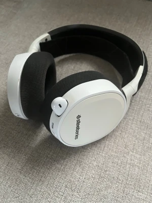 SteelSeries vita trådlösa gaminghörlurar - Säljer ett par vita SteelSeries over-ear gaminghörlurar med svart vadderad bygel och mjuka öronkuddar i tyg. Hörlurarna har inbyggd mikrofon, volymkontroll och flera anslutningsmöjligheter. Perfekta för gaming och långa sessioner tack vare bekväm design. Trådlösa men saknar en sladd, går att köpa billigt på Elgiganten till exempel!