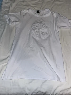 Vit Stone Island t-shirt med logga - Vit t-shirt från Stone Island med stor, broderad logga på bröstet. Klassisk rund hals och korta ärmar. Tillverkad i mjuk bomull som känns skön mot huden. Perfekt för dig som gillar stilrena och trendiga plagg.