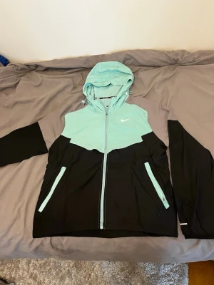 Nike mintgrön och svart vindjacka - Snygg vindjacka från Nike i mintgrönt och svart med grå detaljer på ärmarna. Jackan har huva med dragsko, hel dragkedja och två fickor framtill. Perfekt för blåsiga dagar och sportiga outfits. Liten vit Nike-logga på bröstet.