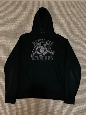 Svart hoodie Reckless Scholars - Svart hoodie med huva från University, prydd med ett stort tryck i silver av en skelettfigur med vingar och texten 'Reckless Scholars' på bröstet. Hoodien har känguruficka och ribbade muddar. Perfekt för dig som gillar streetwear och edgy stil.