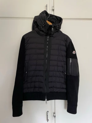 Svart pufferjacka från Moncler - Snygg svart pufferjacka från Moncler med stickade ärmar och vadderad kropp. Jackan har huva med dragsko, tvåvägsdragkedja och fickor med tryckknappar. Moncler-logga på ärmen och extra ficka med dragkedja på armen. Perfekt för kalla vinterdagar.