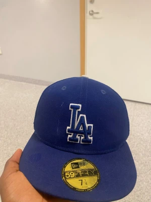 Blå LA Dodgers keps från New Era - Snygg blå keps från New Era med LA Dodgers-logga framtill och mesh på sidorna. Kepsen har broderad 23-patch med kaktus på sidan och klassisk 59FIFTY-form. Perfekt för dig som gillar sportig streetstyle.