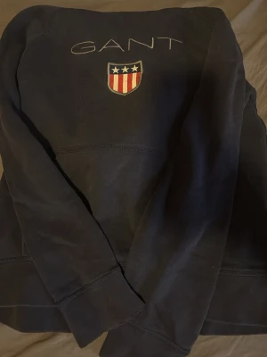 Svart sweatshirt från GANT - Svart sweatshirt från GANT med broderad logga och sköld med stjärnor och ränder på bröstet. Klassisk modell med rund hals och långa ärmar. Perfekt för en avslappnad och stilren look.