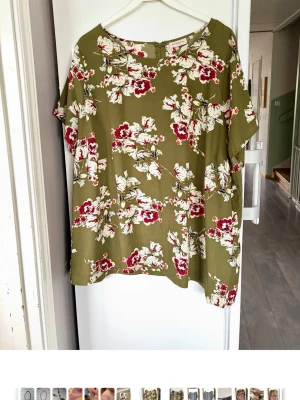 Grön blommig blus med kort ärm plussize - Snygg grön blus med blommigt mönster i vitt och rött. Blusen har en lös passform och korta ärmar, perfekt för varmare dagar. Tillverkad i ett lätt och luftigt material som känns skönt mot huden.