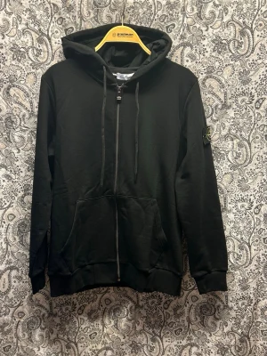 Svart hoodie från Stone Island - Svart hoodie med dragkedja från Stone Island. Klassisk design med huva, dragsnören och känguruficka framtill. Ikonisk logotyp-patch på vänster ärm. Tillverkad i mjukt bomullsmaterial, perfekt för en avslappnad och stilren look.