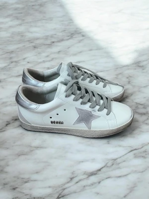 Vita Golden Goose sneakers med silverdetaljer - Snygga vita sneakers från Golden Goose med silverfärgad stjärna på sidan, silvriga detaljer på hälen och grå skosnören. Skorna har en klassisk låg modell och är tillverkade i läder med slitna detaljer på sulan för en cool look.