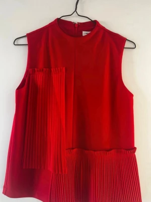 Röd ärmlös topp med plisserade detaljer - Trendig röd topp med plisserade detaljer framtill och nedtill. Ärmlös modell med rund halsringning och dragkedja i ryggen. Perfekt för dig som vill sticka ut med en färgstark look.
