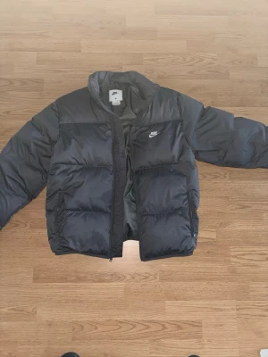 Svart pufferjacka från Nike - Snygg svart pufferjacka från Nike med klassisk logga på bröstet. Jackan har hög krage, dragkedja framtill och två sidofickor. Perfekt för kalla dagar med sin vadderade design och sportiga look. Materialet är glansigt och vattenavvisande.