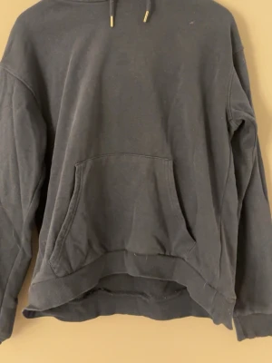 Mörkgrå hoodie med magficka - En enkel och stilren mörkgrå hoodie med huva och dragsnören. Hoodien har en stor magficka framtill och ribbade muddar vid ärmslut och nederkant. Perfekt för en avslappnad och bekväm look. Materialet känns mjukt och skönt mot huden.
