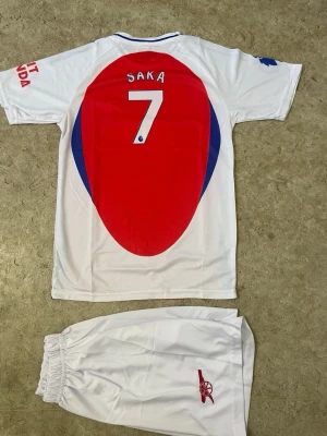 Arsenal fotbollskit Saka #7 - Snyggt Arsenal fotbollskit med Saka #7. Tröjan är röd med vita och blå detaljer, tryck på bröstet och ärmarna samt klubbmärke. Matchande vita shorts med Arsenal-logga. Perfekt för fotbollsträning eller att bära när du hejar på laget.