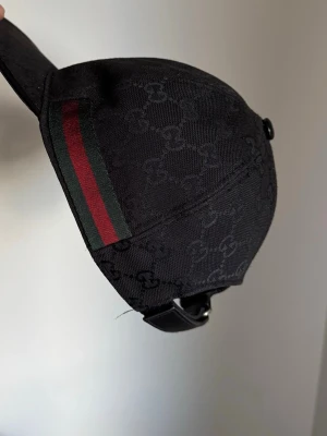 Gucci Keps  - Snygg svart keps från GG med diskret GG-monogram över hela tyget och klassiska gröna och röda ränder på sidan. Justerbar rem baktill i läder ger en lyxig känsla. Perfekt accessoar för en trendig streetstyle-look. Storlek M