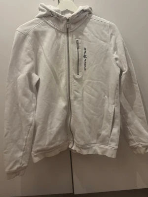 Vit hoodie från Sail Racing - Vit hoodie med dragkedja från Sail Racing. Har huva med dragsko, bröstficka med dragkedja och logga på bröstet. Mjuk bomullskänsla och två sidofickor. Perfekt för en avslappnad och sportig stil.