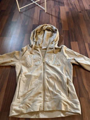 Beige hoodie med dragkedja från USPA - Snygg beige hoodie från USPA med dragkedja framtill och broderad logga på bröstet. Hoodien har huva med snören och två fickor på sidorna. Perfekt för en avslappnad och trendig stil. Materialet känns mjukt och bekvämt.