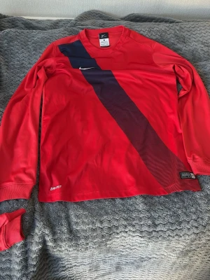 Röd långärmad Nike Dri-FIT tröja  - 147-158 CM - 12-13 år. Snygg röd långärmad sporttröja från Nike med Dri-FIT-teknologi. Tröjan har en diagonal mörkblå rand över framsidan och en vit Nike-logga på bröstet. Perfekt för träning eller fotboll. Tillverkad i lätt och ventilerande material.
