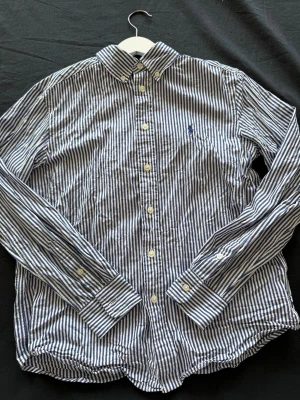 Randig skjorta från Ralph Lauren - Snygg skjorta från Ralph Lauren med blåvita ränder och klassisk krage. Skjortan har långa ärmar, knappar framtill och ikonisk broderad logga på bröstet. Perfekt för en stilren look.
