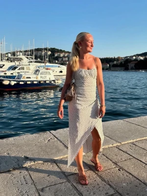 Vit prickig långklänning med slits - Säljer en vit långklänning med svarta prickar och smala axelband. Klänningen har en snygg slits framtill och omlottkänsla nertill. Perfekt för sommaren!☀️