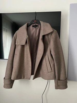 Kort beige kappa från Zara - Stilren kort kappa från Zara i beige ullblandning. Kappan har bred krage, raka fickor och dekorativa manschetter vid ärmsluten. Perfekt för dig som gillar en klassisk och trendig look.