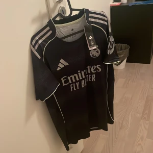 Real Madrid fotbollströja Adidas - Snygg mörkblå Real Madrid fotbollströja från Adidas med vita detaljer och klubbmärke på bröstet. Tröjan har korta ärmar, klassiska Adidas-ränder på axlarna och tryck med 'Emirates Fly Better' på framsidan. Perfekt för alla fotbollsfans! Den är i storlek S. Om ni har frågor kan ni höra av er!