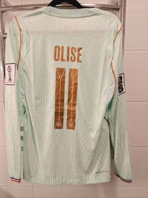 Frankrike Olise matchtröja långärmad spelar version ! - Spelar version lång ärmad VM tröja Ljusblå långärmad fotbollströja från Nike med Frankrikes landslagsmärke, guldiga detaljer och nummer 11 samt namnet Olise på ryggen. Tröjan har FIFA och UEFA märken på ärmarna och är tillverkad i lätt, ventilerande material (Aero-FIT).