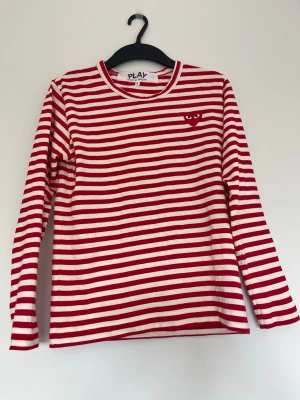 Comme des Garçons långärmad T-shirt  - Säljer en långärmad t-shirt från Comme des Garçons PLAY med röda och vita ränder. Tröjan har ett broderat rött hjärta med ögon på bröstet och är tillverkad i mjuk bomull. Perfekt för en trendig och avslappnad stil. Skulle säga att den passar dig mellan 160 och 175 typ eftersom den funkar både lite stor och lite tajt. Skicket är perfekt och tröjan har använts 1 gång.