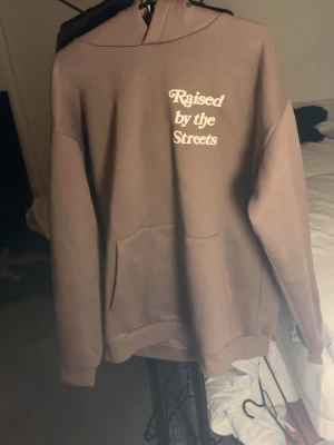 Brun hoodie  - Säljer en brun hoodie från Raised by the Streets med vit text på både bröstet och ryggen. Hoodien har en stor ficka framtill och klassisk huva. Perfekt för dig som gillar streetwear och vill ha en chill look.