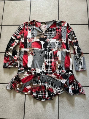 Mönstrad blus med volang från Reese storlek XL - Färgstark blus från Reese med abstrakt mönster i rött, svart, vitt, grått och beige. Blusen har V-ringning, lång ärm och volangdetalj nedtill. Materialet är mjukt och stretchigt, perfekt för en trendig look.