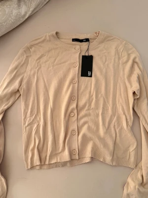 Beige cardigan från 157 - Säljer en stilren beige cardigan från 157 med rund halsringning och knappar framtill. Tröjan har långa ärmar och ribbade muddar vid ärmslut och nederkant. Perfekt att slänga över en topp för en enkel och clean look. Helt oanvänd med prislapp på!