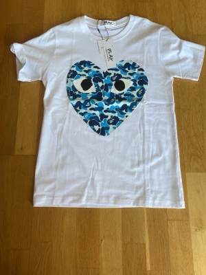 Vit Comme des Garçons Play t-shirt - Vit t-shirt från Comme des Garçons Play med ett stort hjärta på bröstet i blått camouflage-mönster och svarta ögon. Klassisk rund hals och korta ärmar. Perfekt statement-plagg för dig som gillar streetwear och unika prints.