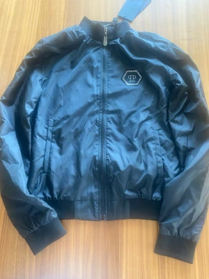Svart bomberjacka från Philipp Plein S tunn sommarjacka - Snygg svart bomberjacka från Philipp Plein med glansig finish och ribbade muddar. Jackan har dragkedja framtill med metalltag och en sexkantig logga med strass på bröstet. Perfekt för dig som gillar streetstyle och exklusiva detaljer. Storlek S. Ny med tag.