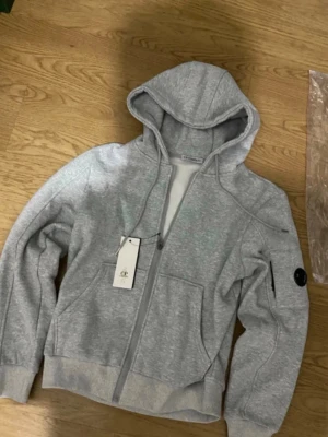 Grå hoodie från C.P. Company - Snygg grå hoodie från C.P. Company med dragkedja framtill, två fickor och klassisk huva. Ärmen har den ikoniska C.P. Company-loggan i svart. Perfekt för en avslappnad och trendig stil. Tillverkad i mjukt material för extra komfort.