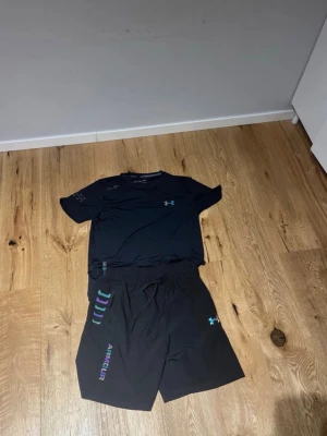 Svart träningsset från Under Armour - Säljer ett svart träningsset från Under Armour med t-shirt och shorts. Båda plaggen har logga och tryck i blått och vitt. Perfekt för gymmet eller löpning. Materialet är lätt och andas, vilket gör det bekvämt under träning.