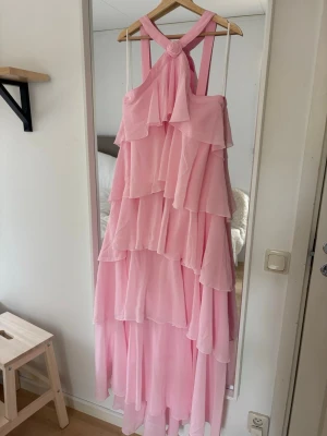 Rosa volangklänning  - Superfin långklänning i ljusrosa med flera lager volanger och breda axelband som korsas i ryggen. Klänningen har omlottkänsla upptill och är tillverkad i ett lätt, flowy material som ger en romantisk vibe. Felicia Wedins kollektion med vero Moda, modellen är VMfelicia. Storlek L.  Aldrig använd med lapp kvar. Nypris 1199 kr, säljer för 600 kr.