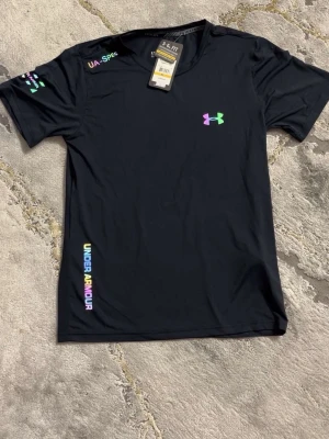 Svart t-shirt från Under Armour - Svart sport-t-shirt från Under Armour med färgglada logotyper och tryckdetaljer i regnbågsfärger. Tillverkad i ett lätt och snabbtorkande funktionsmaterial, perfekt för träning och aktivitet. Klassisk passform med rund hals och korta ärmar.