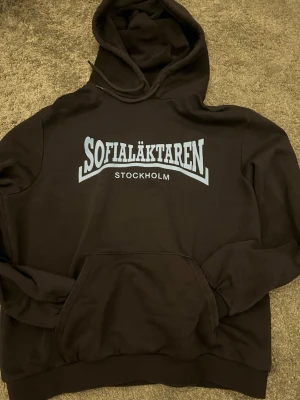 Svart hoodie Sofialäktaren Stockholm - Säljer en svart hoodie från Sofialäktaren med stor vit text på bröstet och 'Stockholm' under. Klassisk känguruficka framtill och justerbar huva med snören. Perfekt för dig som vill representera Stockholm och gillar en avslappnad streetwear-stil. 