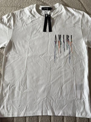Vit AMIRI t-shirt med färgglatt tryck - Vit t-shirt från AMIRI med korta ärmar och rund hals. På bröstet finns ett tryck med AMIRI-loggan och färgglada droppar som ger en unik look. Perfekt för dig som vill sticka ut med en stilren men ändå kreativ design.