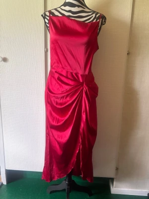 Röd långklänning i satin med drapering - Snygg röd långklänning i glansigt satinmaterial med smala axelband och draperad detalj framtill. Klänningen har omlottkänsla och en elegant slits. Perfekt för dig som vill sticka ut med en klassisk och trendig look.