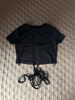 Svart croppad topp från Bershka - Svart croppad topp från Bershka med korta ärmar och långa snören för knytning i midjan. Perfekt till höga jeans eller kjol