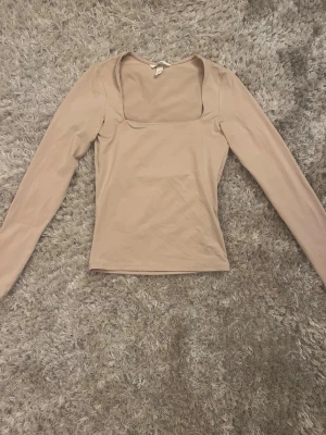 Beige långärmad topp med fyrkantig ringning - Snygg beige topp med lång ärm och fyrkantig ringning. Tillverkad i mjukt material som sitter tight mot kroppen och ger en clean look. Perfekt att styla med jeans eller kjol för en enkel men trendig outfit.