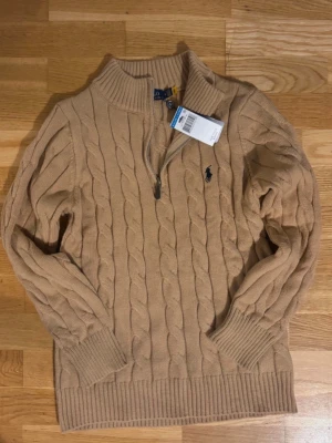 Beige kabelstickad tröja Polo Ralph Lauren - Kabelstickad beige tröja från Polo Ralph Lauren med halv dragkedja och hög krage. Tröjan har ribbade muddar och nederkant samt den klassiska broderade loggan på bröstet. Perfekt för lager-på-lager och snygg till jeans.