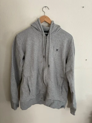 Ljusgrå zip-hoodie från hackett London - Snygg ljusgrå hoodie med dragkedja från Hackett. Två fickor framtill och en liten broderad logga på bröstet. Perfekt för en avslappnad och stilren look. Mjuk bomullsblandning för extra komfort.