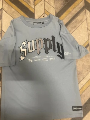 Blå t-shirt från Supply & Demand - Snygg ljusblå t-shirt från Supply & Demand med stort tryck 'supply' i svart och vitt framtill. Klassisk passform med rund hals och korta ärmar. Perfekt för en streetwear-look och enkel att matcha med jeans eller shorts.