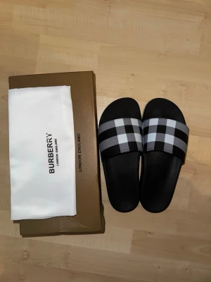 Svarta Burberry tofflor med rutigt band - Säljer ett par svarta tofflor från Burberry med klassiskt rutigt band i svart, vitt och grått. Tofflorna har en stilren och modern design med bred rem över foten och är tillverkade i syntetmaterial. Perfekta för en trendig look.