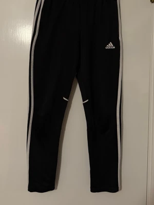 Svarta Adidas träningsbyxor med ränder - Svarta träningsbyxor från Adidas med klassiska vita ränder längs sidorna och Adidas-logga på låret. Byxorna har smal passform och resår i midjan. Perfekta för träning eller sportiga outfits.