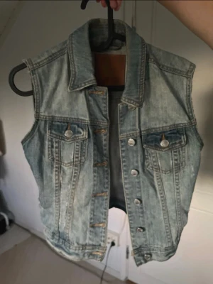 Ärmlös ljusblå jeansväst - Snygg ärmlös jeansjacka i ljusblå denim med klassisk krage och knappar framtill. Två bröstfickor med lock och silverfärgade knappar.
