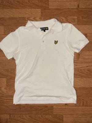 Vit pikétröja från Lyle & Scott - Klassisk vit pikétröja från Lyle & Scott med korta ärmar och gul logga på bröstet. Tröjan har krage och knäppning med två knappar framtill. Tillverkad i bomull för en skön och luftig känsla.