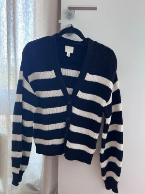 Randig svartvit kofta från H&M - Snygg stickad kofta från H&M i svart och vitt med breda horisontella ränder. Koftan har v-ringning, tre knappar framtill och ribbade muddar vid ärmslut och nederkant. Perfekt för en trendig och avslappnad look.
