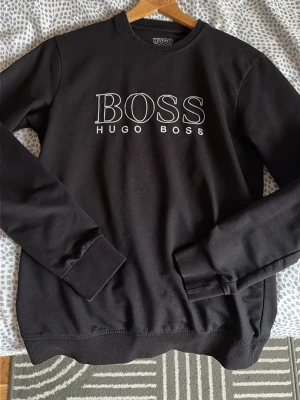 Svart sweatshirt från Hugo Boss - Svart sweatshirt med rund hals och vit BOSS-logga framtill. Tröjan har långa ärmar, ribbade muddar och nederkant. Perfekt för en stilren och avslappnad look.