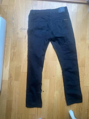 Svarta jeans från Nudie Jeans - Säljer ett par svarta jeans från Nudie Jeans med klassisk femficksdesign och raka ben. Jeansen har diskret logga på bakfickan och är tillverkade i ett slitstarkt bomullstyg. Perfekta för dig som gillar stilrena och tidlösa plagg. Dessutom använd en gång !